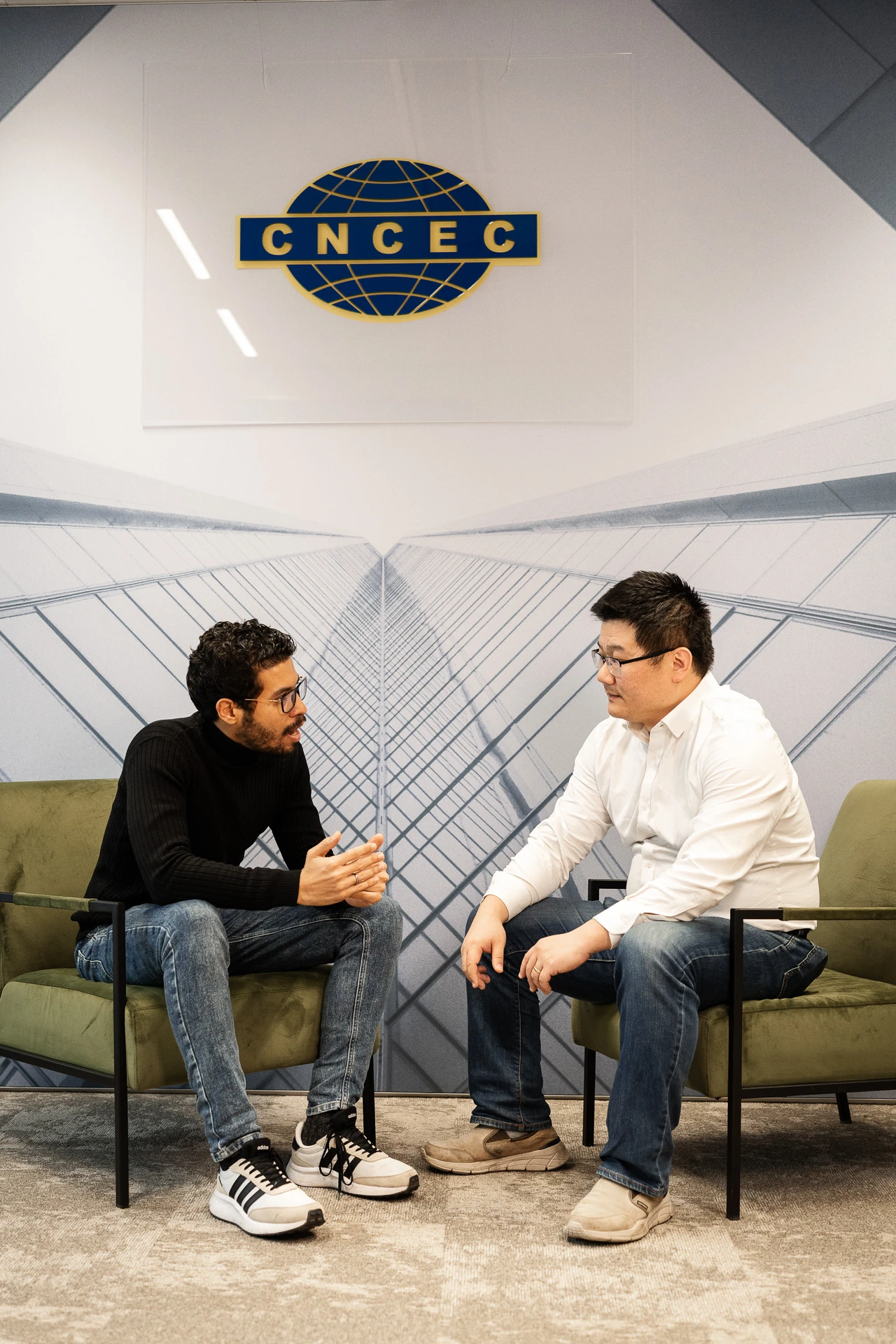 2 men sitting on 2 green armchairs conversing under the CNCEC logo. 2 men sitting on 2 green armchairs conversing under the CNCEC logo.