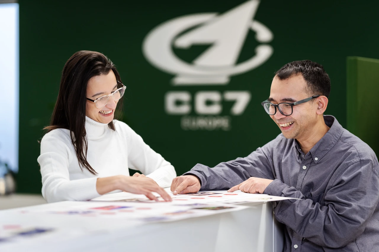 1 woman and 1 man smiling and conversing standing in front of a CC7 Europe logo. 1 woman and 1 man smiling and conversing standing in front of a CC7 Europe logo.