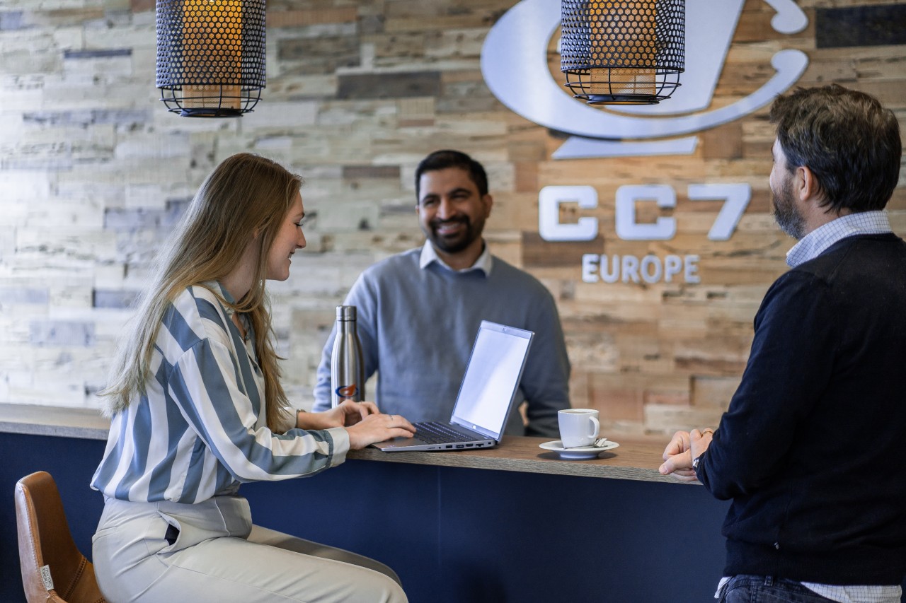 3 people conversing while smiling and working at the laptop with the CC7 Europe logo on the background. 3 people conversing while smiling and working at the laptop with the CC7 Europe logo on the background.
